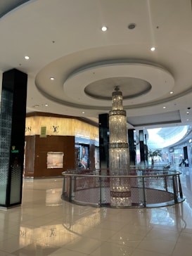 A luxurious indoor shopping mall featuring high-end stores such as Louis Vuitton. The mall has glossy tiled floors, a large central chandelier descending from the ceiling, and plenty of ambient lighting. The decor is modern and sophisticated, with clean lines and a spacious layout.