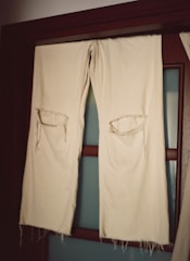 A pair of off-white fabric trousers with large torn holes at the knees hangs over a wooden rod in front of a frosted glass door. The fabric appears worn and frayed at the edges.