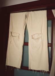 A pair of off-white fabric trousers with large torn holes at the knees hangs over a wooden rod in front of a frosted glass door. The fabric appears worn and frayed at the edges.