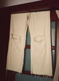 A pair of off-white fabric trousers with large torn holes at the knees hangs over a wooden rod in front of a frosted glass door. The fabric appears worn and frayed at the edges.