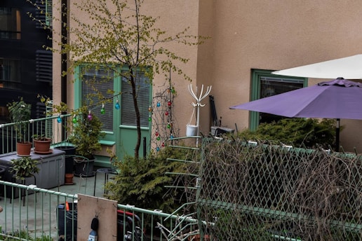 A small, cozy balcony with potted plants and colorful string lights. There is a green metal railing along the edge, and a purple patio umbrella provides shade. The beige building wall features a green door and window. There are some shrubs and a small tree, adding to the greenery.
