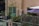 A small, cozy balcony with potted plants and colorful string lights. There is a green metal railing along the edge, and a purple patio umbrella provides shade. The beige building wall features a green door and window. There are some shrubs and a small tree, adding to the greenery.