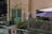 A small, cozy balcony with potted plants and colorful string lights. There is a green metal railing along the edge, and a purple patio umbrella provides shade. The beige building wall features a green door and window. There are some shrubs and a small tree, adding to the greenery.