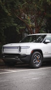 A modern electric truck is parked on a street with lush greenery surrounding it. The vehicle is white with sleek design lines and features distinctive headlights. The background includes a tree with dense foliage.