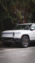 A modern electric truck is parked on a street with lush greenery surrounding it. The vehicle is white with sleek design lines and features distinctive headlights. The background includes a tree with dense foliage.