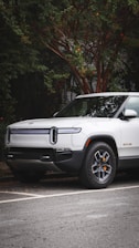 A modern electric truck is parked on a street with lush greenery surrounding it. The vehicle is white with sleek design lines and features distinctive headlights. The background includes a tree with dense foliage.