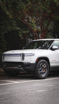 A modern electric truck is parked on a street with lush greenery surrounding it. The vehicle is white with sleek design lines and features distinctive headlights. The background includes a tree with dense foliage.