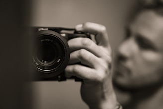 A close-up image of a person holding a camera in their hand, with a focus on the camera lens. The background and the person are blurred, highlighting the camera as the main subject. The photo is in black and white, giving it a classic and timeless feel.
