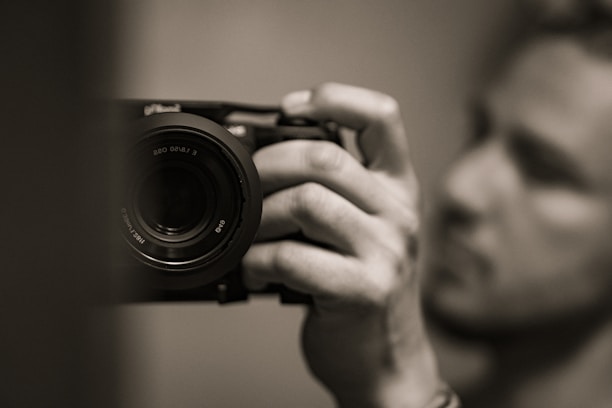 A close-up image of a person holding a camera in their hand, with a focus on the camera lens. The background and the person are blurred, highlighting the camera as the main subject. The photo is in black and white, giving it a classic and timeless feel.