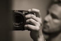 A close-up image of a person holding a camera in their hand, with a focus on the camera lens. The background and the person are blurred, highlighting the camera as the main subject. The photo is in black and white, giving it a classic and timeless feel.