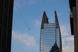 A tall, modern skyscraper with a unique split design at the top, featuring glass windows and structural supports. There is a large logo displayed near the top with the text 'King & Spalding'. The sky is blue with scattered clouds, and there is a thin wire crossing the image diagonally with birds perched on it. Nearby buildings partially frame the scene on both sides.