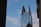 A tall, modern skyscraper with a unique split design at the top, featuring glass windows and structural supports. There is a large logo displayed near the top with the text 'King & Spalding'. The sky is blue with scattered clouds, and there is a thin wire crossing the image diagonally with birds perched on it. Nearby buildings partially frame the scene on both sides.