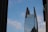 A tall, modern skyscraper with a unique split design at the top, featuring glass windows and structural supports. There is a large logo displayed near the top with the text 'King & Spalding'. The sky is blue with scattered clouds, and there is a thin wire crossing the image diagonally with birds perched on it. Nearby buildings partially frame the scene on both sides.