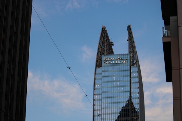 A tall, modern skyscraper with a unique split design at the top, featuring glass windows and structural supports. There is a large logo displayed near the top with the text 'King & Spalding'. The sky is blue with scattered clouds, and there is a thin wire crossing the image diagonally with birds perched on it. Nearby buildings partially frame the scene on both sides.