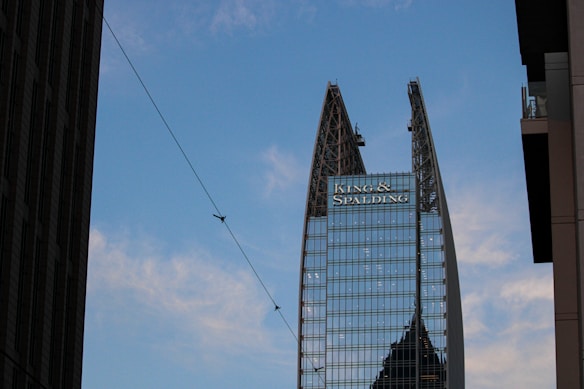 A tall, modern skyscraper with a unique split design at the top, featuring glass windows and structural supports. There is a large logo displayed near the top with the text 'King & Spalding'. The sky is blue with scattered clouds, and there is a thin wire crossing the image diagonally with birds perched on it. Nearby buildings partially frame the scene on both sides.
