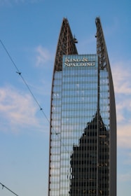 A modern skyscraper with a unique angular design features reflective glass panels. The name 'King & Spalding' is prominently displayed at the top, and another building’s silhouette is visible in the reflection. The blue sky and some thin clouds are visible in the background.