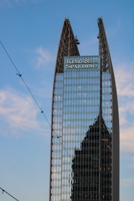 A modern skyscraper with a unique angular design features reflective glass panels. The name 'King & Spalding' is prominently displayed at the top, and another building’s silhouette is visible in the reflection. The blue sky and some thin clouds are visible in the background.