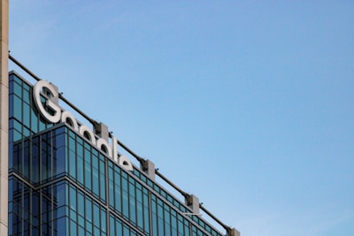 The image depicts a modern glass building with the word 'Google' prominently displayed near the top. The building features reflective glass windows and a clear blue sky serves as the backdrop.