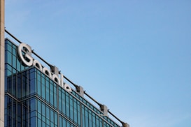 The image depicts a modern glass building with the word 'Google' prominently displayed near the top. The building features reflective glass windows and a clear blue sky serves as the backdrop.