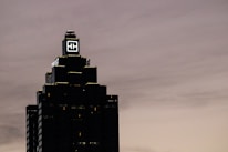 A tall, dark skyscraper is silhouetted against a cloudy sky. The building has a prominent logo with the letters 'HH' illuminated near the top. The outline of the structure is highlighted by lights on each tier, and there is a sign that reads 'Truist' along one side.