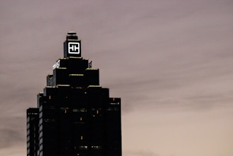 A tall, dark skyscraper is silhouetted against a cloudy sky. The building has a prominent logo with the letters 'HH' illuminated near the top. The outline of the structure is highlighted by lights on each tier, and there is a sign that reads 'Truist' along one side.