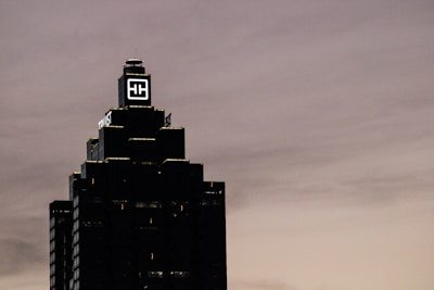 A tall, dark skyscraper is silhouetted against a cloudy sky. The building has a prominent logo with the letters 'HH' illuminated near the top. The outline of the structure is highlighted by lights on each tier, and there is a sign that reads 'Truist' along one side.