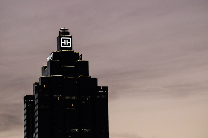 A tall, dark skyscraper is silhouetted against a cloudy sky. The building has a prominent logo with the letters 'HH' illuminated near the top. The outline of the structure is highlighted by lights on each tier, and there is a sign that reads 'Truist' along one side.