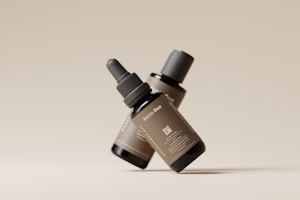Two dark glass dropper bottles with labels are arranged in a minimalist style against a neutral beige background. The bottles are positioned at an angle, and one prominently displays the text 'Bottle Duo' along with a QR code and additional small print details.