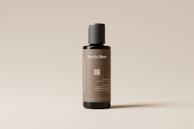 A sleek, cylindrical bottle with a minimalist design stands against a light neutral background. The label on the bottle reads 'Bottle Duo' in white text, and there is a QR code printed below it. The bottle's color is a muted brown with a black cap, and additional text is displayed vertically along the side.