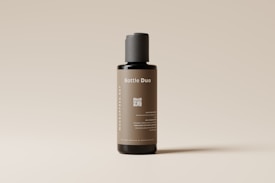 A sleek, cylindrical bottle with a minimalist design stands against a light neutral background. The label on the bottle reads 'Bottle Duo' in white text, and there is a QR code printed below it. The bottle's color is a muted brown with a black cap, and additional text is displayed vertically along the side.