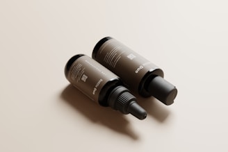 Two brown cosmetic bottles lie on a smooth, light beige surface. One bottle has a screw top with a dropper, while the other has a pump dispenser. The labels display text and a QR code.