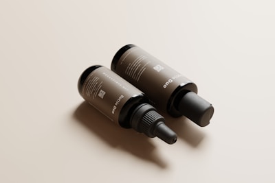 Two brown cosmetic bottles lie on a smooth, light beige surface. One bottle has a screw top with a dropper, while the other has a pump dispenser. The labels display text and a QR code.