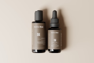 Two bottles are placed side by side on a neutral beige background. The larger bottle has a pump dispenser, while the smaller bottle features a dropper. Labels on both bottles read 'Bottle Duo' with detailed text below, including a QR code. The bottles have a sleek and minimalist design.
