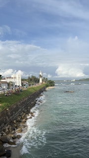 A scenic coastal area featuring a lighthouse, historical buildings, and a rocky shoreline. The ocean is calm with gentle waves, and the sky is partly cloudy with blue skies peeking through. People are gathered near the grassy area, enjoying the view.