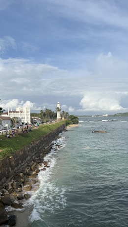 A scenic coastal area featuring a lighthouse, historical buildings, and a rocky shoreline. The ocean is calm with gentle waves, and the sky is partly cloudy with blue skies peeking through. People are gathered near the grassy area, enjoying the view.