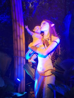 A figure with blue skin and distinct facial features holds a child under vibrant lighting in a jungle-like setting. The light casts a dramatic blue and orange glow, highlighting the textured leaves and surrounding foliage.