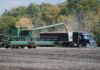 A large green combine harvester unloads grain into a truck trailer in a harvested field, set against a backdrop of dense green and autumn-colored trees.