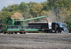 A large green combine harvester unloads grain into a truck trailer in a harvested field, set against a backdrop of dense green and autumn-colored trees.