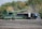 A large green combine harvester unloads grain into a truck trailer in a harvested field, set against a backdrop of dense green and autumn-colored trees.