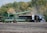 A large green combine harvester unloads grain into a truck trailer in a harvested field, set against a backdrop of dense green and autumn-colored trees.
