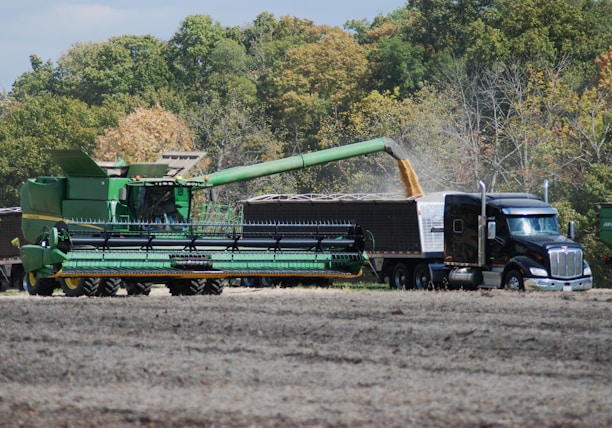 A large green combine harvester unloads grain into a truck trailer in a harvested field, set against a backdrop of dense green and autumn-colored trees.