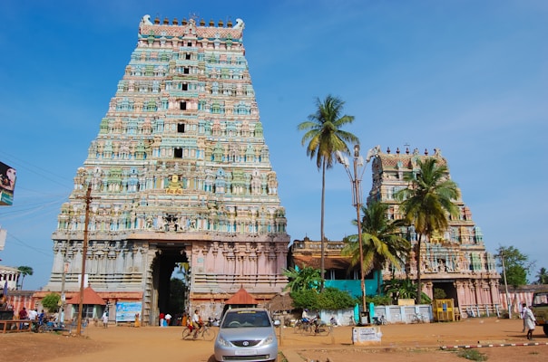 A large and intricately designed temple entrance with two towering gopurams adorned with colorful carvings and sculptures. The taller tower is on the left, featuring detailed artwork, including deities and ornamental motifs. Surrounding the temple are palm trees, adding to the serene atmosphere. People and vehicles are seen in the area, suggesting activity around the temple.