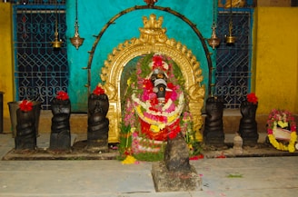 A religious altar featuring a Ganesha idol adorned with vibrant garlands made of flowers like red, white, and yellow. The idol is set against a backdrop of a golden ornate arch with intricate designs. Black stone serpent statues with red flowers placed on top are lined on either side of the idol. The setting includes hanging brass ornaments and a background of teal and yellow walls with patterned grillwork.