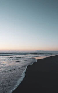 A tranquil beach scene at dusk with soft waves and a muted blue sky blending into the horizon.