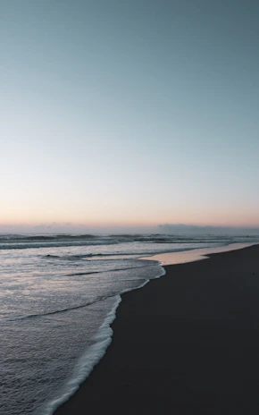 A tranquil beach scene at dusk with soft waves and a muted blue sky blending into the horizon.