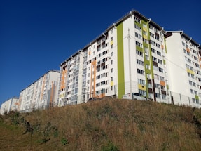 Several modern apartment buildings with multiple floors and colorful facades, including green, white, and orange panels. The buildings are situated on a sloped grassy area under a clear blue sky.