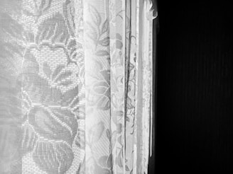 Intricately patterned lace curtains with floral designs hang in front of a darkened, likely black, background. The lace displays detailed, delicate flowers and leaves, allowing some light to filter through, creating a textured, soft appearance.