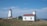 A picturesque coastal scene featuring a white lighthouse with a red roof, surrounded by a few small buildings including one with a matching red roof. The setting is peaceful, with a grassy foreground leading to a rocky shoreline, and the sea extending to the horizon under a blue sky with light clouds.