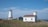 A picturesque coastal scene featuring a white lighthouse with a red roof, surrounded by a few small buildings including one with a matching red roof. The setting is peaceful, with a grassy foreground leading to a rocky shoreline, and the sea extending to the horizon under a blue sky with light clouds.
