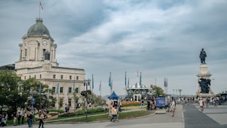 A historic building with a clock tower stands prominently, surrounded by people walking and engaging in leisure activities. A large statue is visible in the background, and flags are waving around a small pavilion amid flowerbeds.